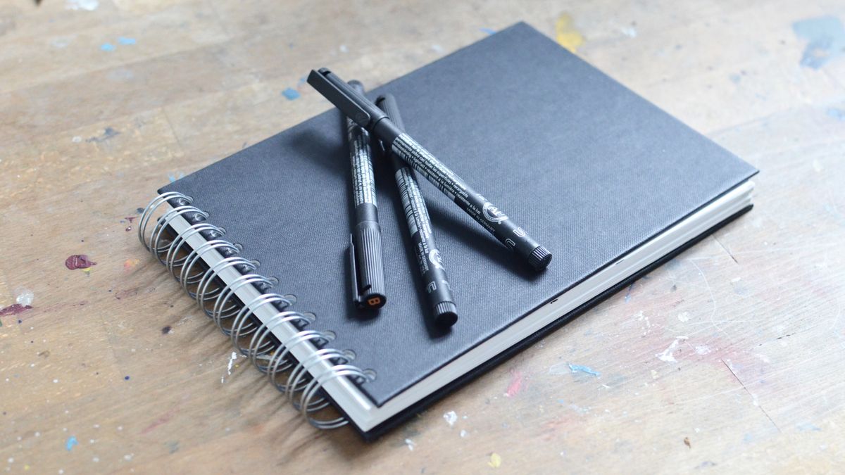 Drawing pens beside a spiral sketchbook on a table