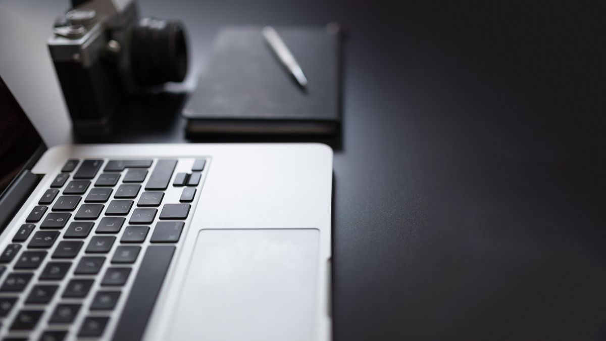 A camera, laptop and notebook arranged on a dark desk